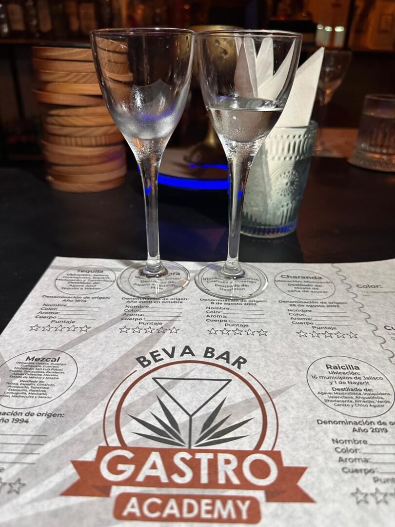 Dos copas medianas sobre un mantel de papel que dice: Beva Bar Gastro Academy.